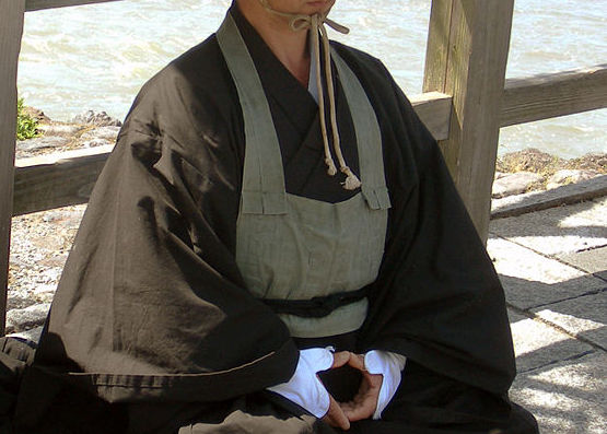 A zen monk sitting outside on concrete wearing a straw hat and tan colored rakusu.